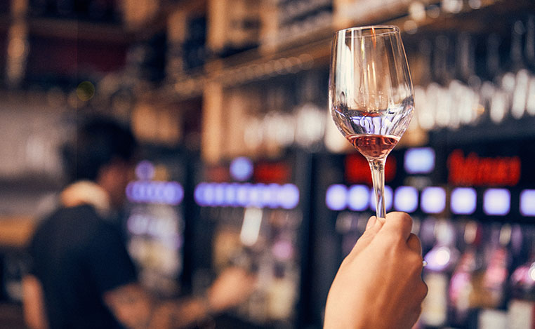 Photo de la main et du verre d'une personne qui lève son verre de vin rouge pour l'observer (première étape de la dégustation) devant la partie libre-service du 5 Wine Bar (arrière plan flou)
